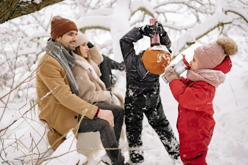 Family enjoying a snowy winter day outdoors, drinking from mugs in warm clothes.